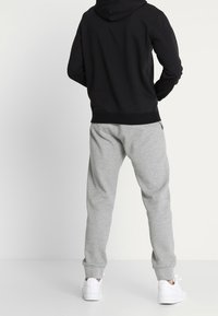 Persona vista da dietro, che indossa un hoodie nero, pantaloni felpati grigio chiaro e scarpe da tennis bianche, mentre si trova davanti a uno sfondo chiaro e uniforme.