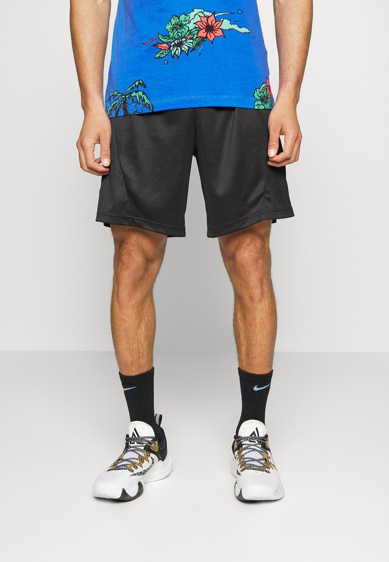 Homme portant une chemise bleue à motifs floraux, un short de sport noir, des chaussettes noires Nike et des baskets blanches, debout devant un fond uni.
