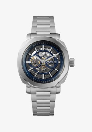 Stålur med skeleton urskive, blå detaljer og børstet finish. Uret har et sølvfarvet metalarmbånd og synlige bevægelseskomponenter.