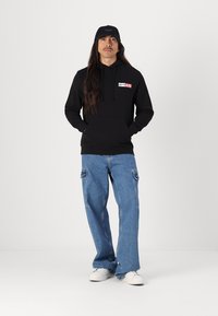 Sort hættejakke med logo, blå cargo jeans med sidelommer, hvide sneakers og en matchende sort bøttehat. Lige snit.