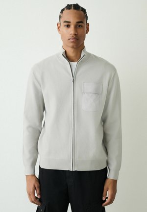 Light gray zip-up jacket in a soft textured fabric, featuring a high collar and a chest pocket. Black pants complete the outfit.
