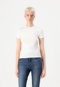 Hvit kortermet t-skjorte laget av glatt stoff, med rund hals og tettsittende design, kombinert med mørkeblå jeans. Ingen mønstre eller detaljer.