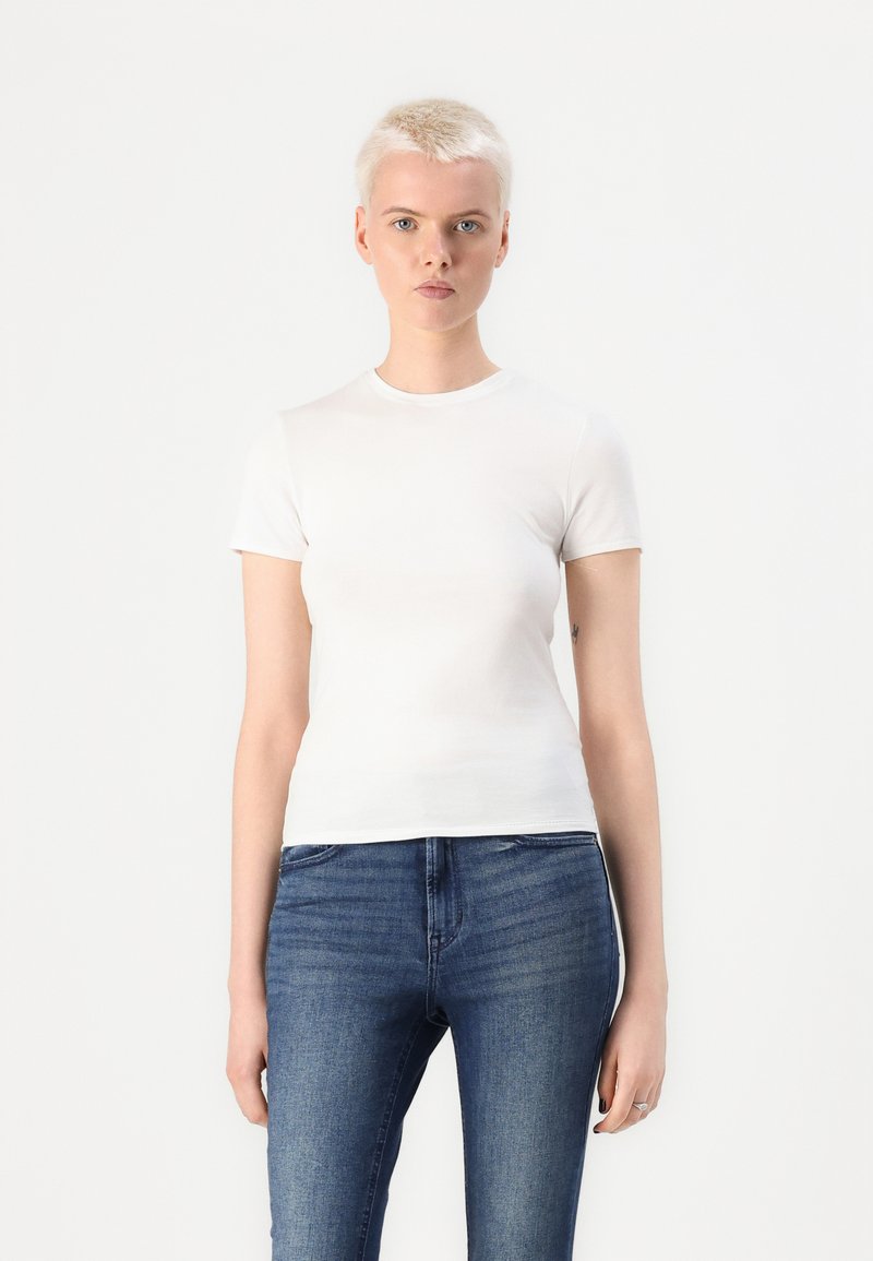 Hvit kortermet t-skjorte laget av glatt stoff, med rund hals og tettsittende design, kombinert med mørkeblå jeans. Ingen mønstre eller detaljer.