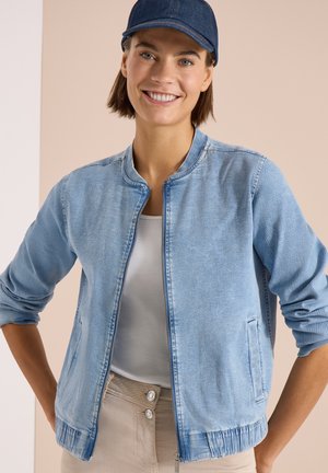 Młoda kobieta uśmiechająca się, ubrana w niebieską kurtkę jeansową, białą koszulę, beżowe spodnie i ciemnoniebieską czapkę baseballówkę, pozująca na neutralnym tle.