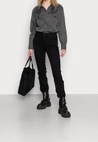 Schwarze, hoch taillierte Jeans, graues Hemd mit Knöpfen und zwei Brusttaschen sowie klobige schwarze Kampfstiefel. Das Model trägt eine schwarze Tragetasche.