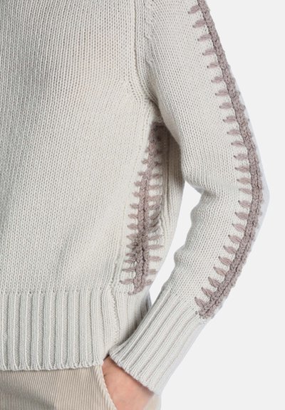 Pull en maille crème avec des surpiqûres décoratives taupe le long de la manche et de la couture latérale, porté avec la main dans la poche d’un pantalon beige.