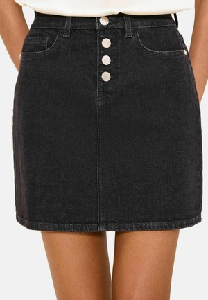 Mini-jupe en jean noire taille haute avec quatre boutons argentés sur le devant et poches avant, portée avec un haut blanc rentré.