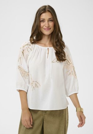 Žena nosí bielu blúzku s výšivkou motýľov a voľnými rukávmi, skombinovanú s olivovo zelenými nohavicami a zlatým náramkom na zápästie.