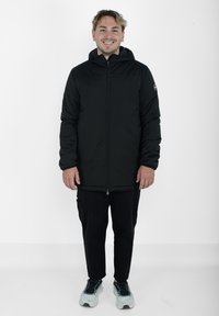 Sort hættetrøje-pufferjakke med en frontlynlås, der har en glat tekstur og et logo-patch på ærmet, kombineret med sorte bukser og sneakers.