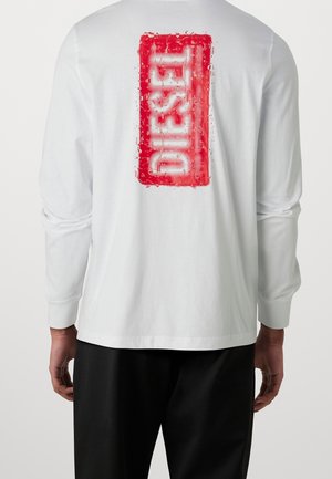 Person wearing a white long-sleeve shirt with a vertical red rectangular Diesel logo on the back and black pants.