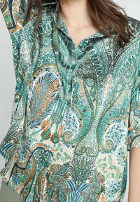Chemise à manches courtes dans un tissu léger, arborant un motif floral paisley multicolore vibrant. Elle possède un col et une fermeture à boutons, avec des accents verts.