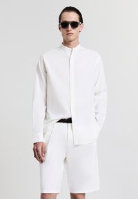 Camicia bianca a maniche lunghe con colletto alla mandarino, tessuto testurizzato, chiusura con bottoni, abbinata a pantaloni corti bianchi e una cintura intrecciata. Occhiali da sole neri.