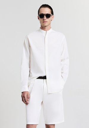 Weißes langärmliges Hemd mit Stehkragen, strukturierter Stoff, knöpfbarer Vorderseite, kombiniert mit weißen Shorts und einem geflochtenen Gürtel. Schwarze Sonnenbrille.