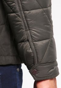 Manche d'une veste matelassée vert foncé avec poignet boutonné portée sur un bras en jean bleu.