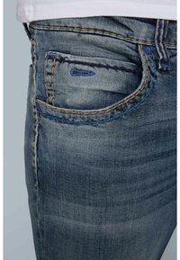 Vaqueros de denim en azul desgastado con costuras en contraste beige y azul. Cuenta con un bolsillo frontal de forma redondeada y un cierre clásico de botón.