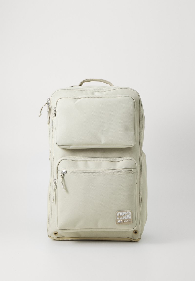 Light beige backpack made from durable fabric, featuring two zippered front pockets, a large compartment, and a top handle for easy carrying.