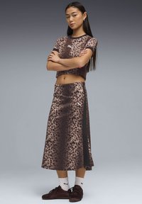 Ung kvinde med langt hår iført leopardmønstret kort top og matchende midi nederdel, hvide Puma-strømper og brune sneakers, med armene krydset.