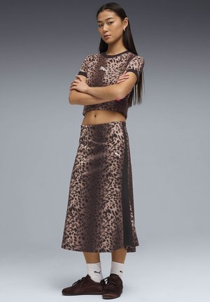 Jonge vrouw met lang haar, gekleed in een cropped top met luipaardprint en een bijpassende midi-rok, witte Puma-sokken en bruine sneakers, met gekruiste armen.