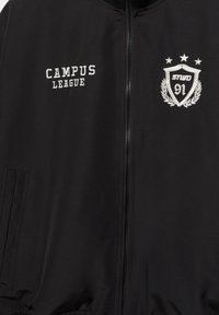 Zwarte jas met rits, voorzien van "CAMPUS LEAGUE" in witte borduring, een schildontwerp met "91" en lauwerkransdetails. Gestructureerde stof, twee zijzakken.