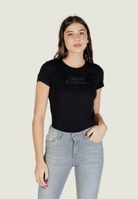Jeune femme aux longs cheveux bruns portant un t-shirt noir Armani Exchange et un jean gris clair, main dans la poche, fond neutre.