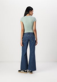 Camiseta justa verde-clara, jeans de cintura alta azuis e flare, e ténis pretos com riscas brancas. Tecido suave e pernas largas.