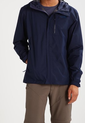 Homme portant une veste à capuche bleu marine avec poche poitrine zippée et pantalon marron, debout avec une main dans la poche.