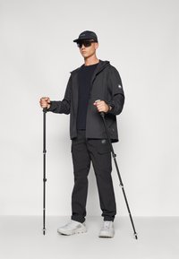 Svart vattentät jacka med justerbar huva, svarta cargobyxor, vita vandringsskor och vandringsstavar. Tillbehör inkluderar en svart keps och solglasögon.