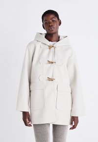 Un manteau en mélange de laine beige clair avec une capuche, des fermetures à bascule et des poches avant, présentant des épaules structurées et une silhouette décontractée.