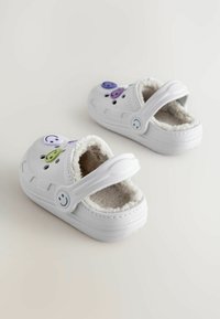Weiße Clogs mit weichem Futter aus Fell, ausgestattet mit bunten Smiley-Gesicht-Charms in Lila und Grün. Verstellbarer Riemen für einen sicheren Sitz.