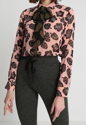 Vrouw draagt een roze blouse met zwarte blad- en bloemprints en een zwart-olijfgroene strik, gecombineerd met hooggelede geribbelde donkergrijze broek.
