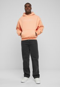 Sudadera melocotón con un bolsillo frontal y logo en blanco, combinada con pantalones gris oscuro y zapatillas blancas. Tejido suave, corte relajado.