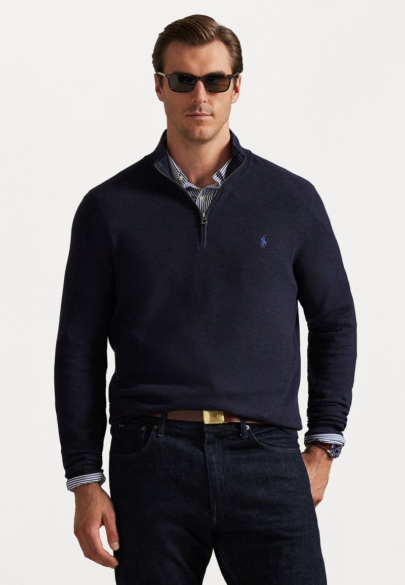 Σκούρο ναυτικό ζακέτα με φερμουάρ, κατασκευασμένη από υφασμάτινη ύλη, φορεμένη πάνω από πουκάμισο με μπλε και λευκές ρίγες, συνδυασμένη με σκούρα τζιν και γυαλιά ηλίου.