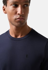 Mørkeblå T-shirt lavet af glat stof, med en diskret rund halsudskæring og et lille, præget bogstav 'B' på brystet.