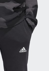 Felpa nera mimetica e pantaloni jogger grigio scuro con logo bianco Adidas. La felpa ha un collo rotondo e polsini a costine; i pantaloni jogger presentano una tasca.