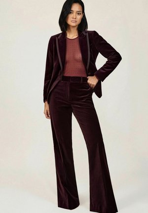 Costume en velours bourgogne avec une veste ajustée et un pantalon évasé. Haut transparent bordeaux avec de petits pois. Revers détaillés et coupe slim.