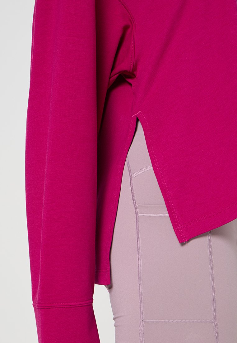 Top de manga larga en fucsia con un corte relajado, que presenta aberturas laterales y una textura suave, combinado con pantalones deportivos de color morado claro.