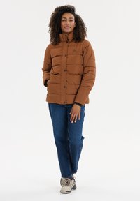 Brun pufferjakke med høj krave, vandret syningsmønster og sorte trykknapper, parret med blå jeans og lyse sneakers.