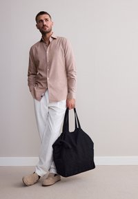 Light pink long-sleeve shirt, white linen trousers, beige slip-on shoes, holding a black tote bag with two handles. Neutral background.
