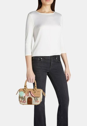 Donna con top bianco e jeans neri che tiene una borsa beige a forma di casa decorata con fiori e un palloncino a forma di cuore.