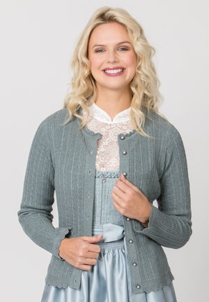 Blonde Frau lächelt, trägt grauen strukturierten knöpfbaren Cardigan über weißer Spitzenbluse und hellblauem Satinsrock, passt den Cardigan an.