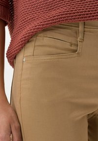 Enfoque cercano de una persona con pantalones color beige y un suéter de punto texturado en color óxido, mostrando el bolsillo delantero y la mano descansando al lado.