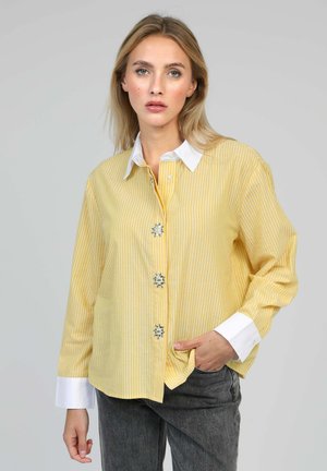 Femme aux longs cheveux blonds portant une chemise rayée jaune et blanche avec des boutons décoratifs et un jean gris foncé, main dans la poche.