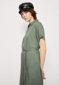 Abito verde con bottoni e maniche corte, con una vita legata, realizzato in un tessuto fluido. Presenta una silhouette rilassata e linee pulite.