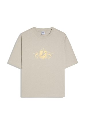 T-shirt beige à manches courtes avec un motif de blason jaune et le texte « Achieve Anything » sur le devant, avec des étoiles et des éléments décoratifs.