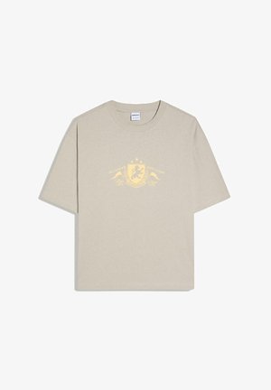 T-shirt beige à manches courtes avec un motif de blason jaune et le texte « Achieve Anything » sur le devant, avec des étoiles et des éléments décoratifs.