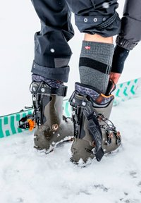 Skischuhe aus dunkelgrauem Kunststoff mit schwarzen und orangefarbenen Akzenten, ausgestattet mit Schnallen und Schnürung. Schneebedeckter Boden und teilweise sichtbarer Ski.