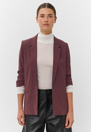 Femme aux cheveux coiffés en arrière portant un col roulé blanc, un blazer prune avec les manches retroussées et un pantalon en cuir noir, debout devant un fond uni.