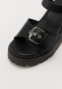 Černé kožené sandály na platformě se širokým páskem, který má stříbrnou sponu a texturovanou gumovou podrážku. Hladký povrch nahoře.