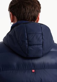 Marinblå pufferjacka med avtagbar huva, med kviltat mönster, kontrasterande svart foder och en liten röd logotyp på baksidan.