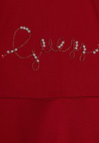Rotes Sweatshirt mit dem Wort "Guess" in Kursivschrift, umrandet mit metallischem Faden und verziert mit weißen Kunstperlen.